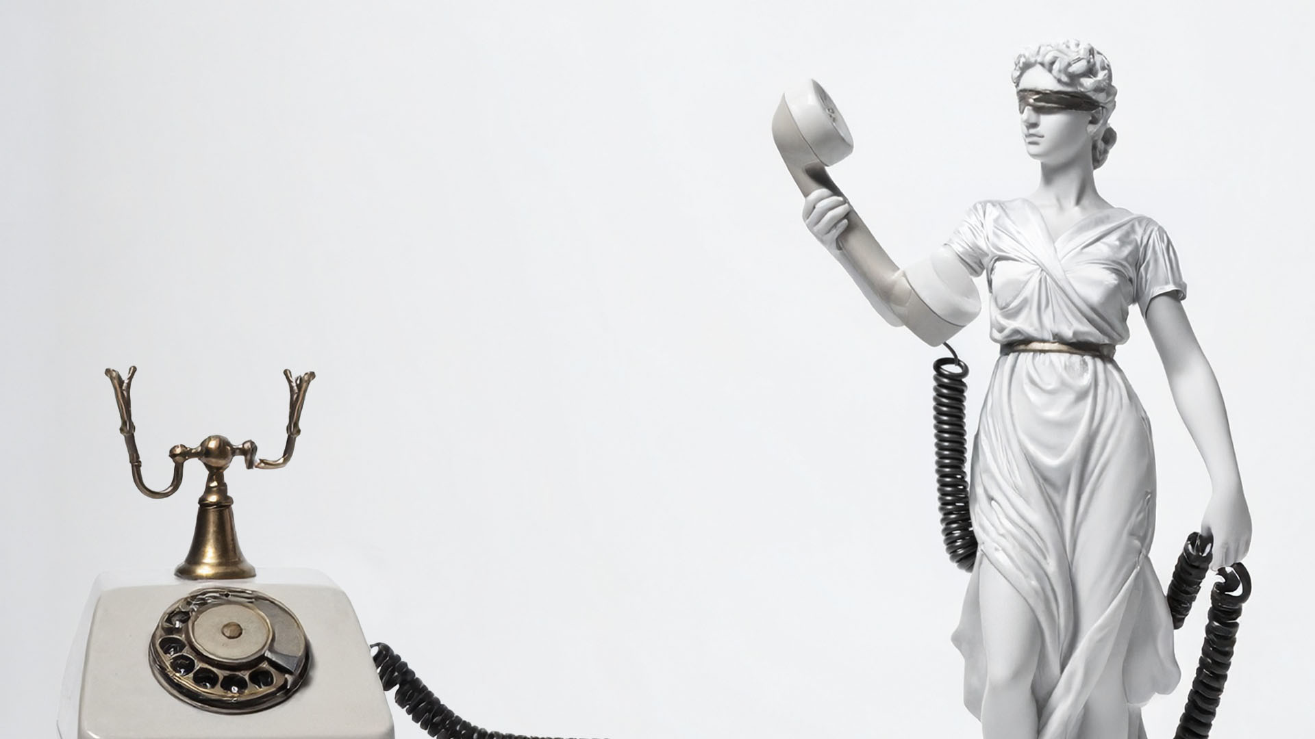 Blindfolded Lady Justice statue holding a telephone receiver beside a vintage rotary phone.