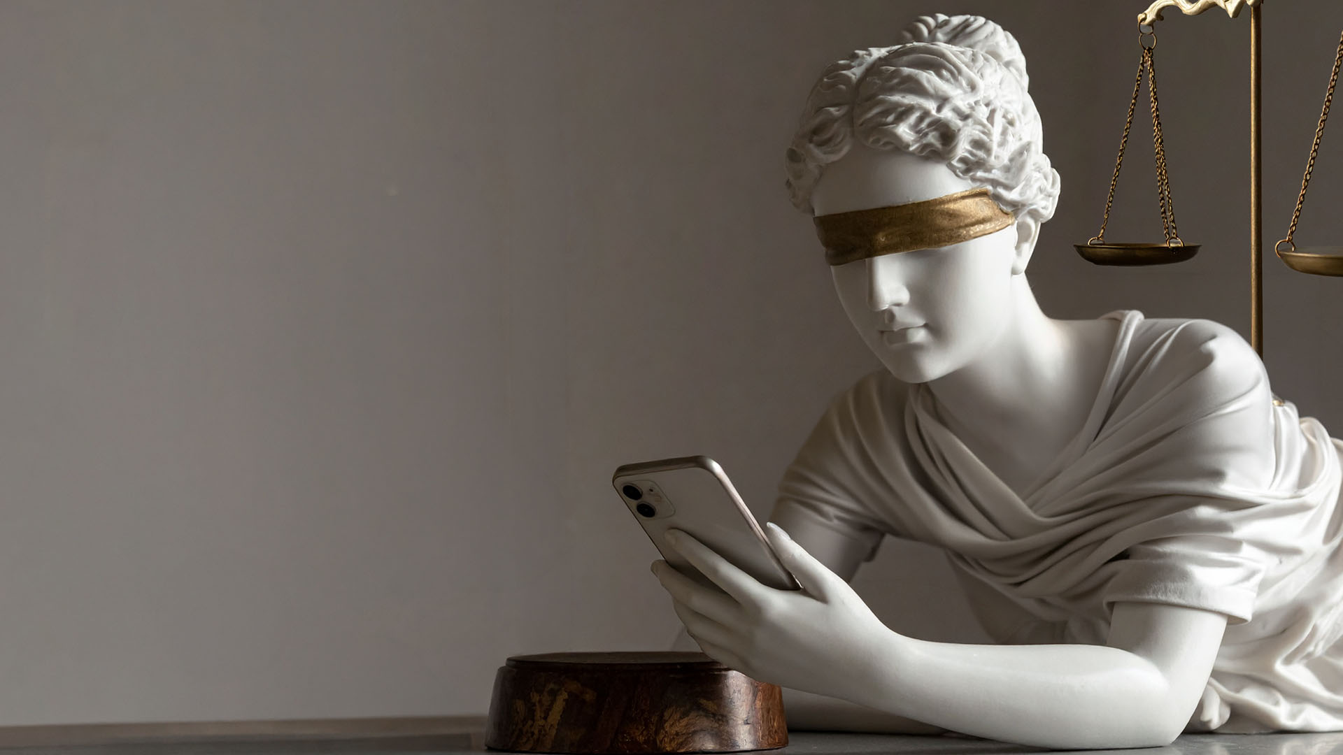 White classical statue of a blindfolded woman holding a smartphone, with brass scales of justice behind her.